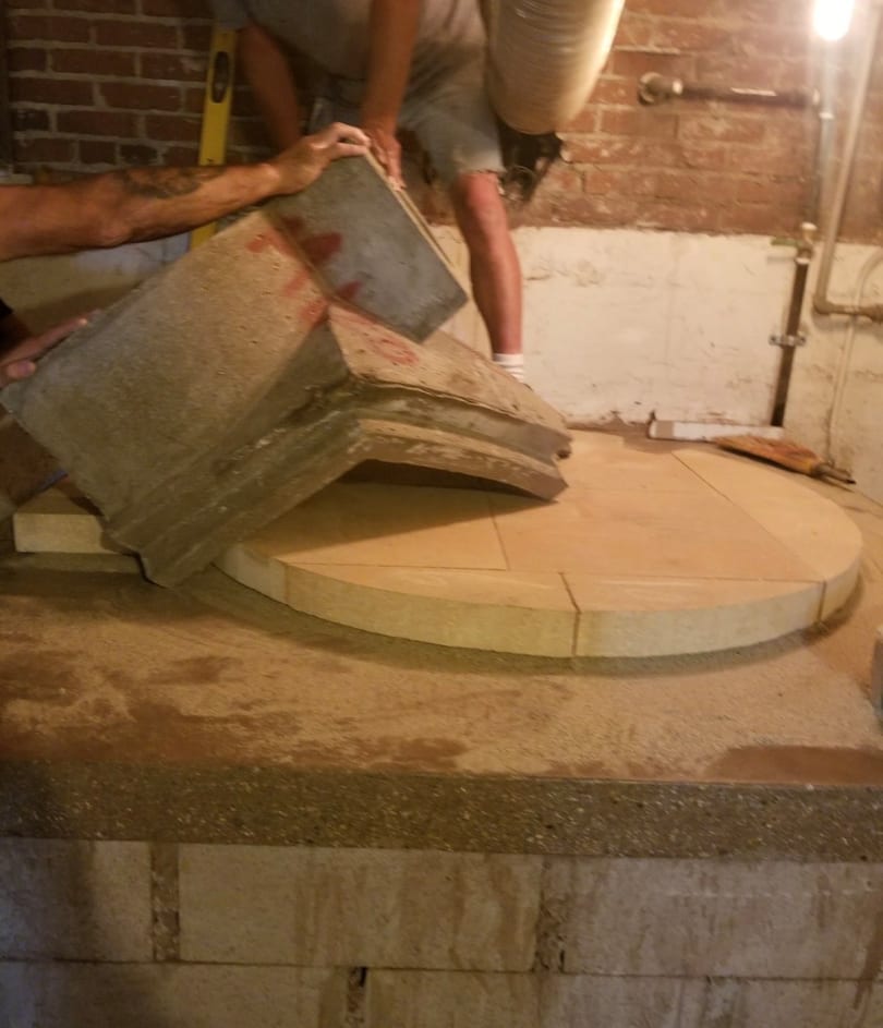 Two people construct a circular oven base with large stone pieces against a brick wall.