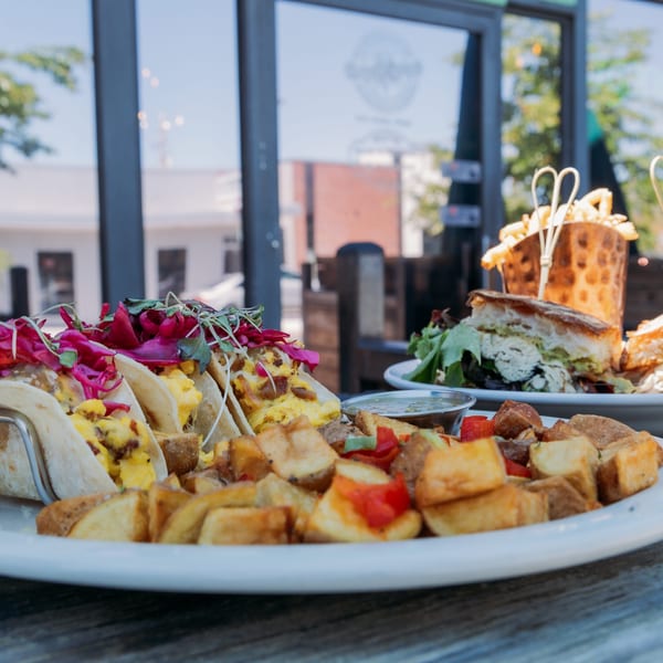 Plates of breakfast tacos, home fries, and a sandwich with fries on an outdoor restaurant patio.