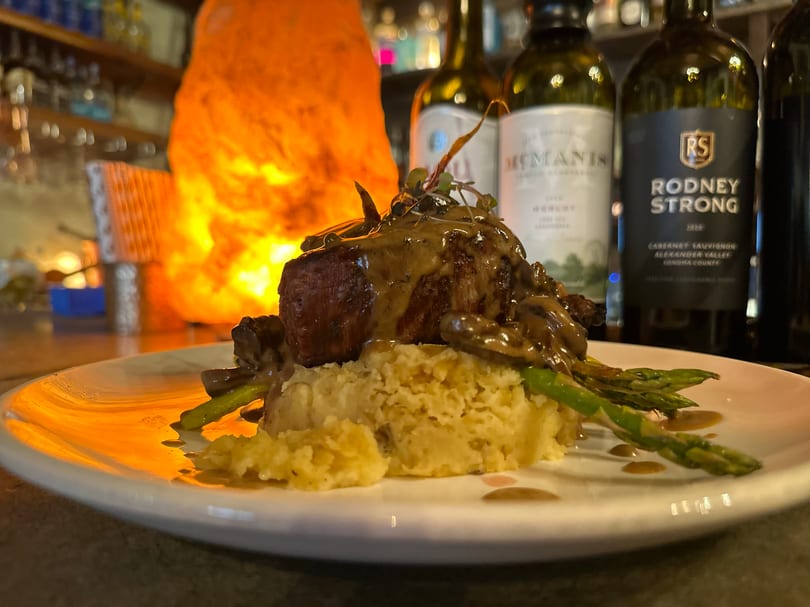 Plated steak with mushroom gravy, mashed potatoes, and asparagus, with wine bottles and a warm lamp behind.