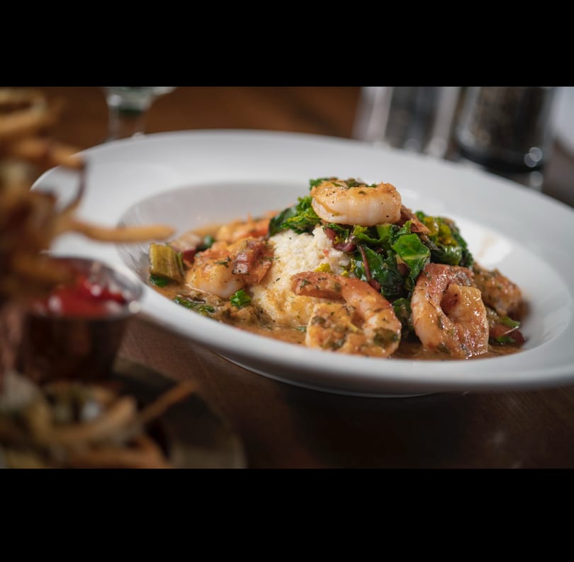 White bowl of shrimp and grits topped with greens and a savory sauce.