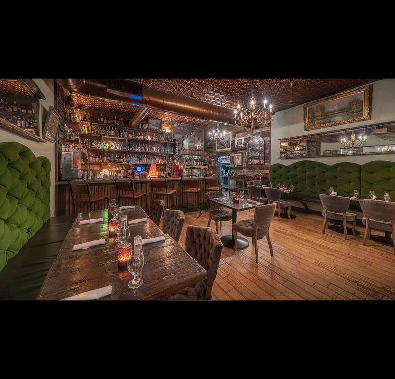 Cozy restaurant interior featuring a well-stocked bar, wooden tables, and plush green booth seating.