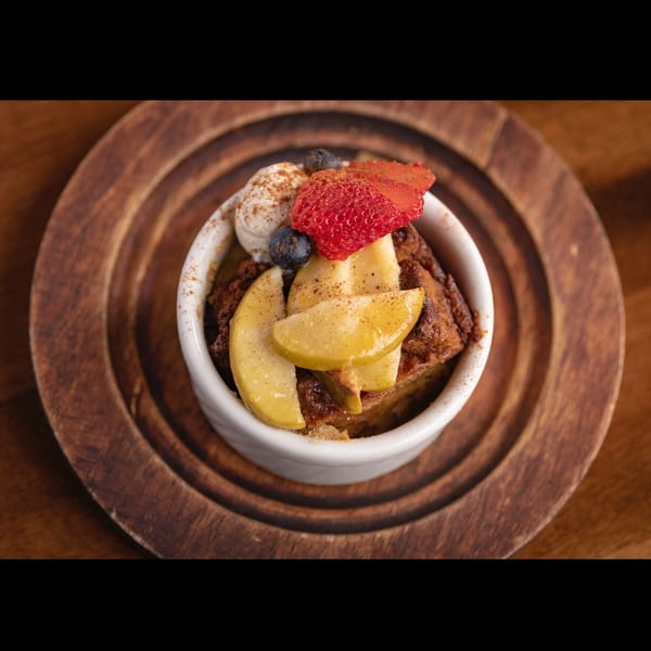 Bread pudding topped with whipped cream, strawberries, blueberries, and apple slices on a wooden plate.