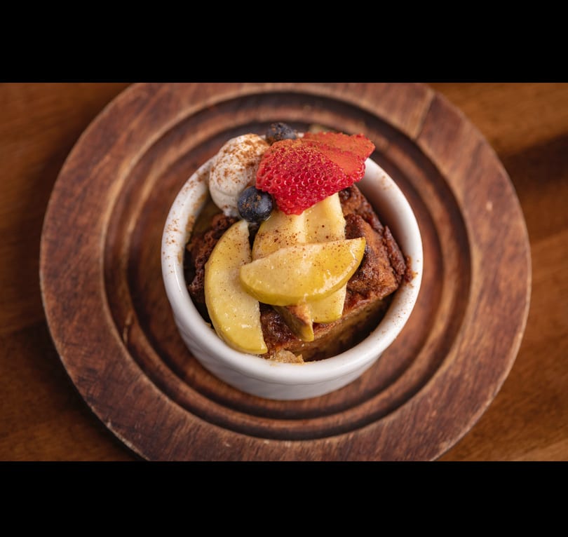 Bread pudding topped with whipped cream, strawberries, blueberries, and apple slices on a wooden plate.